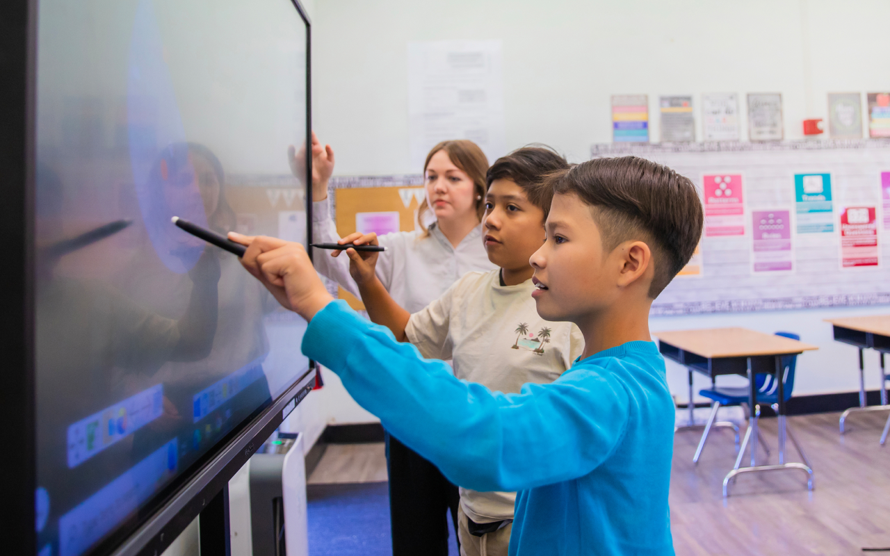 Two students use styluses to interact with a ViewSonic ViewBoard in a classroom while an educator observes. The classroom features colorful posters and empty desks in the background.