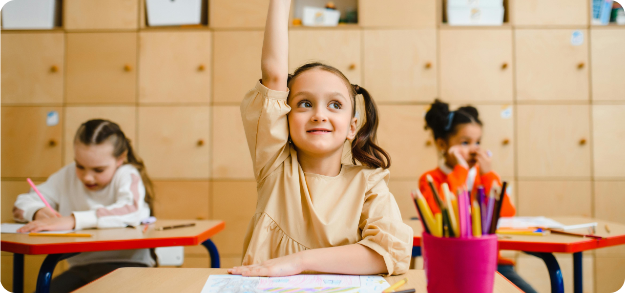 A student raises a hand eagerly during a first day of school activity, with two others working nearby in a warm, well-organized classroom.