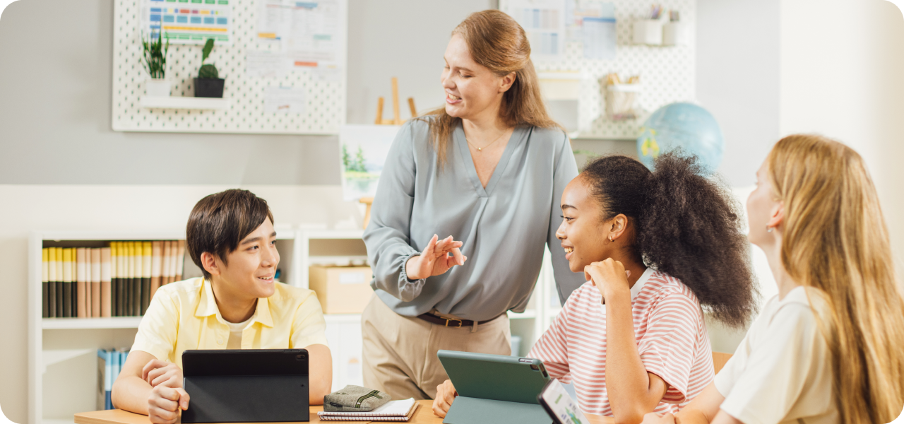 An instructor leads a small group first day of school activity with three students using a laptop and printed materials in a bright, modern classroom.