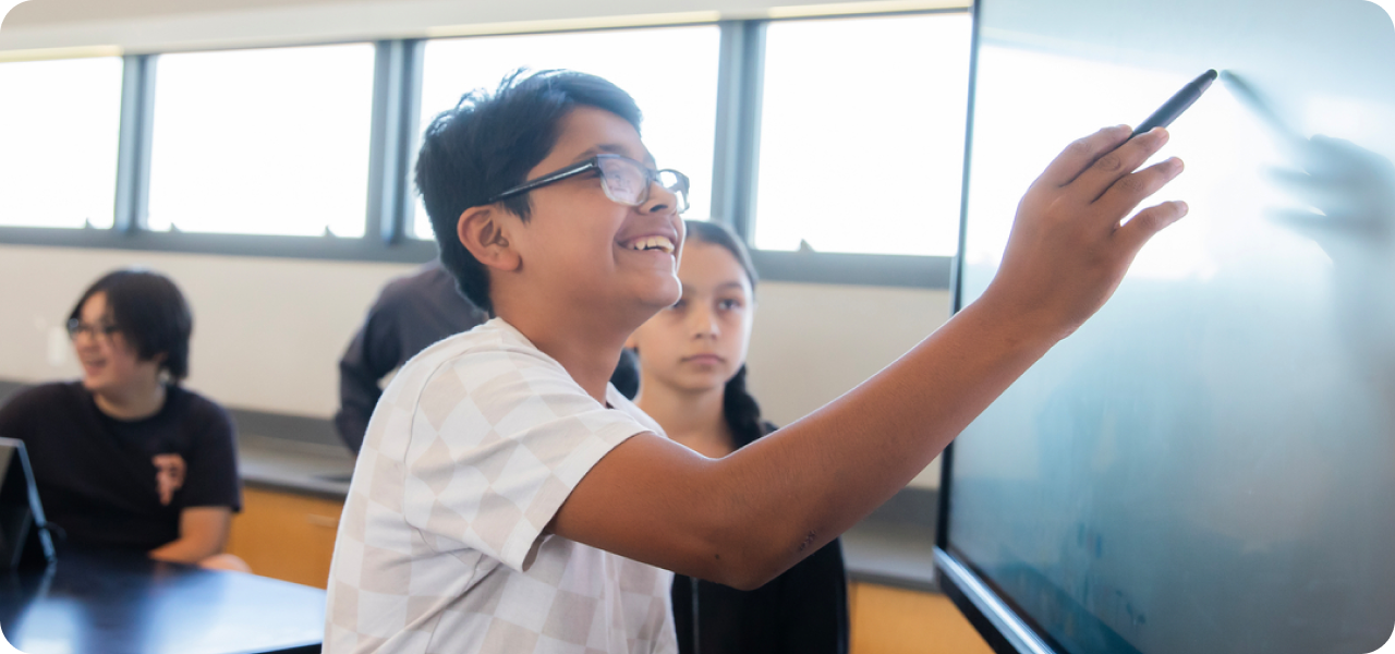 A student explains something at the whiteboard during a first day of school activity, gesturing with a marker while classmates look on from their seats.
