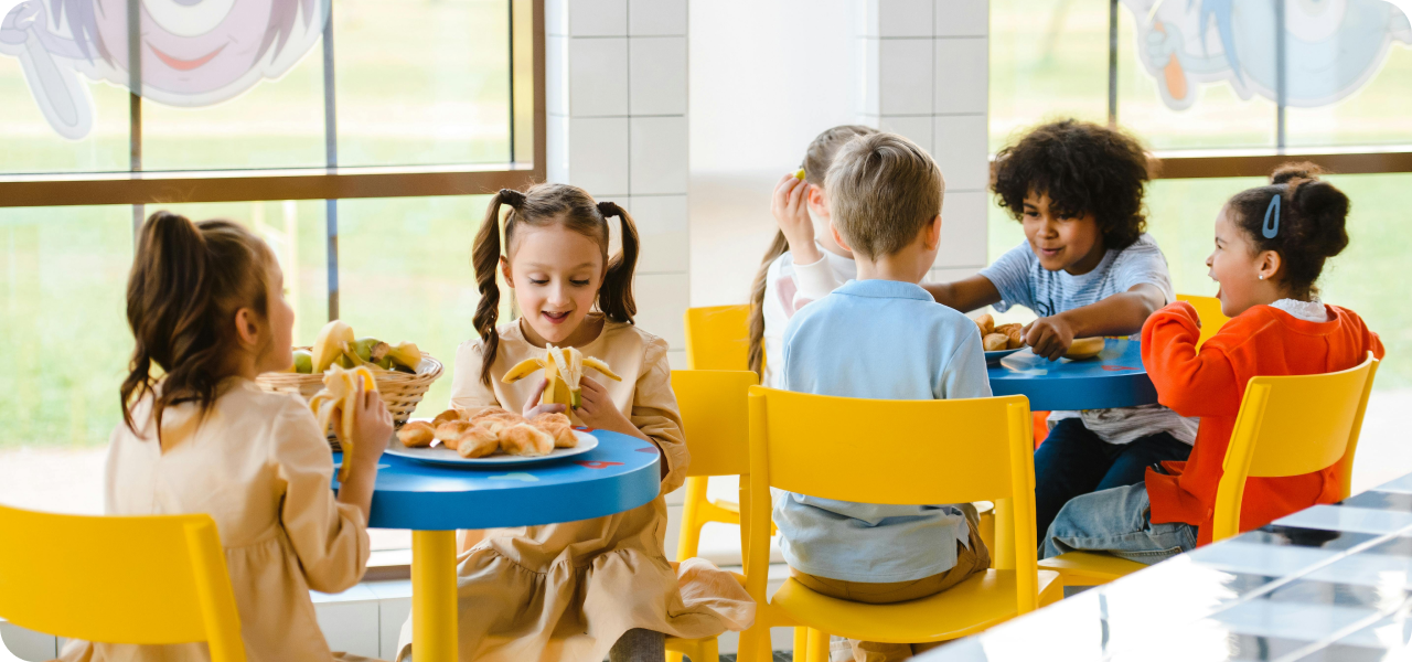A group of elementary students sit around a colorful lunch table, chatting and laughing while enjoying their meals in a sunny classroom.