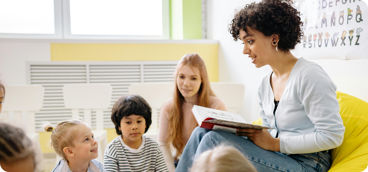 A young teacher reads a book aloud to an engaged group of young students sitting together on the classroom floor, with an alphabet chart visible in the background.