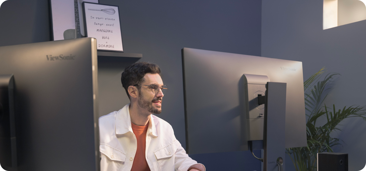A man wearing glasses and a white jacket works at a desk with two ViewSonic ColorPro VP2788-5K monitors in a modern, softly lit workspace. A plant is visible in the background, along with wall art and shelving.