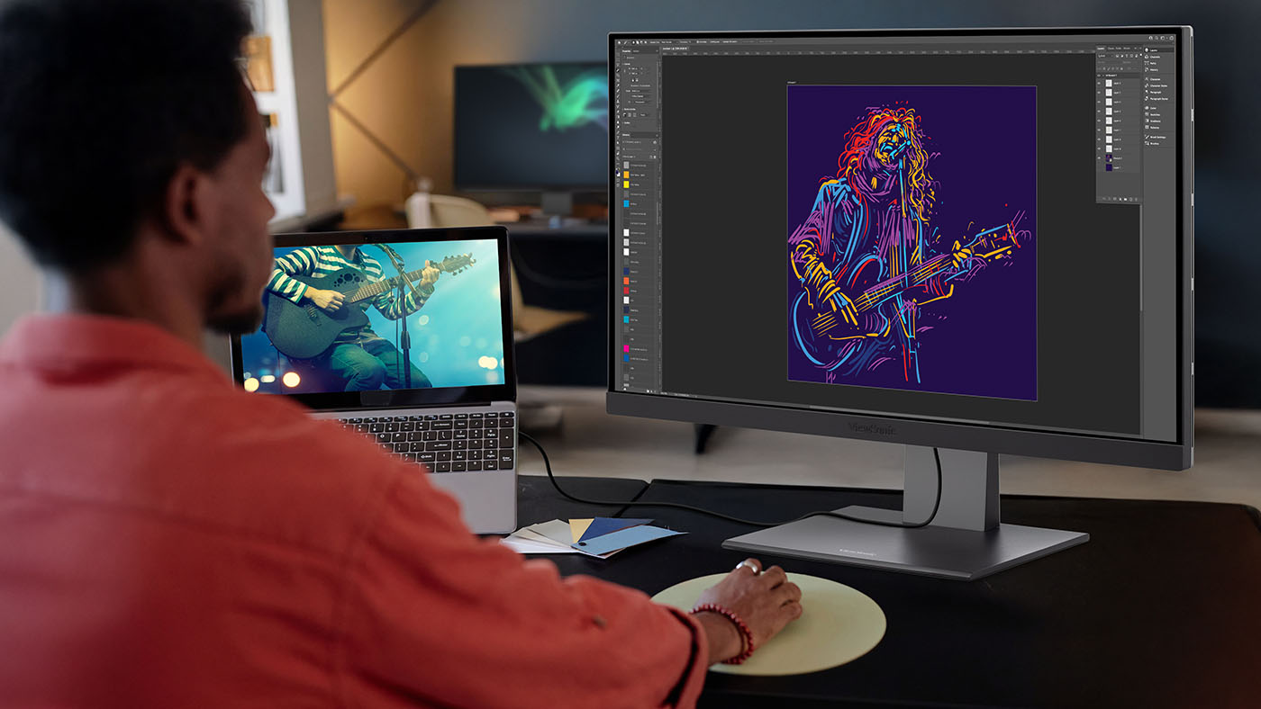 A man in a red shirt looking at a monitor showing a colorful image of a man playing guitar.