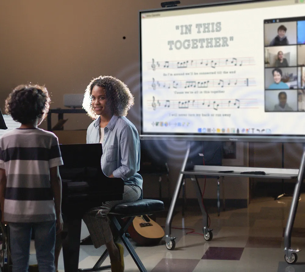 a music teacher seated at a piano teaching a student sheet music that is displayed on the screen in the background.