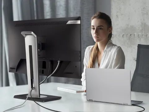 a woman sitting at a monitor connected to a laptop