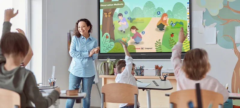a teacher stands in front of a classroom with a viewboard on the wall while students raise their hands