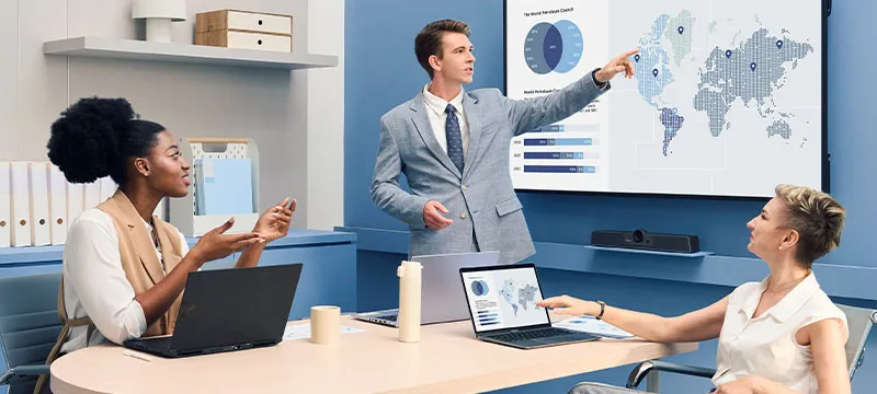 man standing and pointing to a CDE31 display with 2 women coworkers sitting at a table with laptops also looking at the CDE31