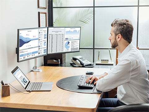 Man working at desk with side-by-side dual business monitors displaying complex finance spreadsheets and analytics software charts