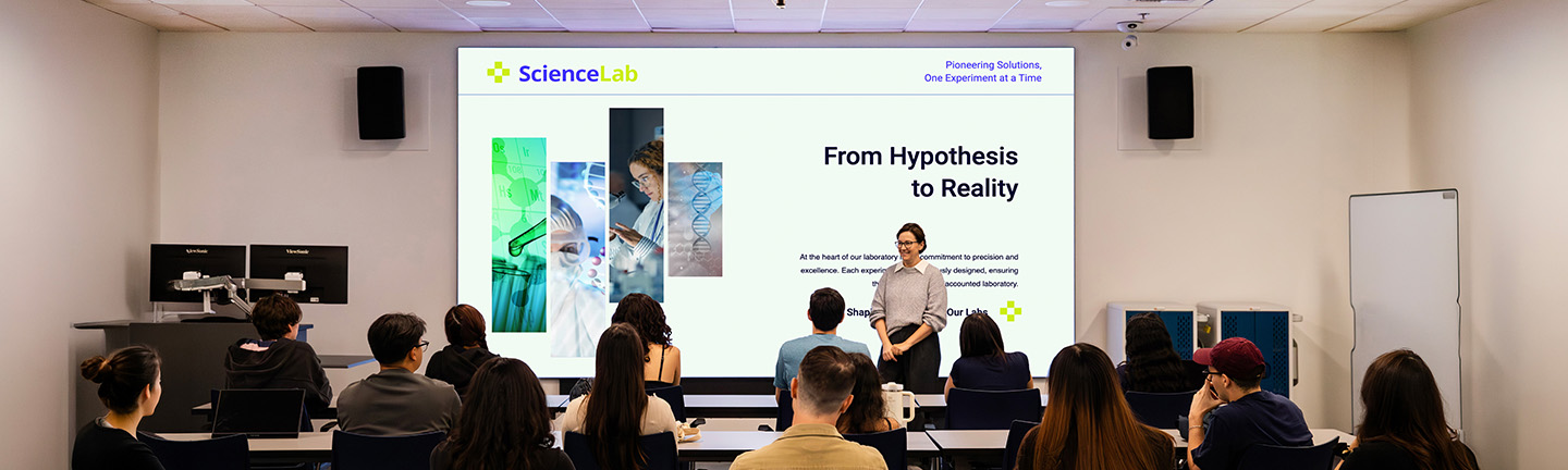 Instructor at the front of a higher ed classroom standing in front of a large format screen
