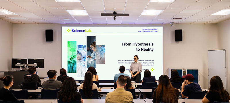 Instructor at the front of a higher ed classroom standing in front of a large format screen
