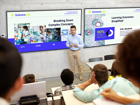 a professor stands at the front of a lecture hall in front of two large displays