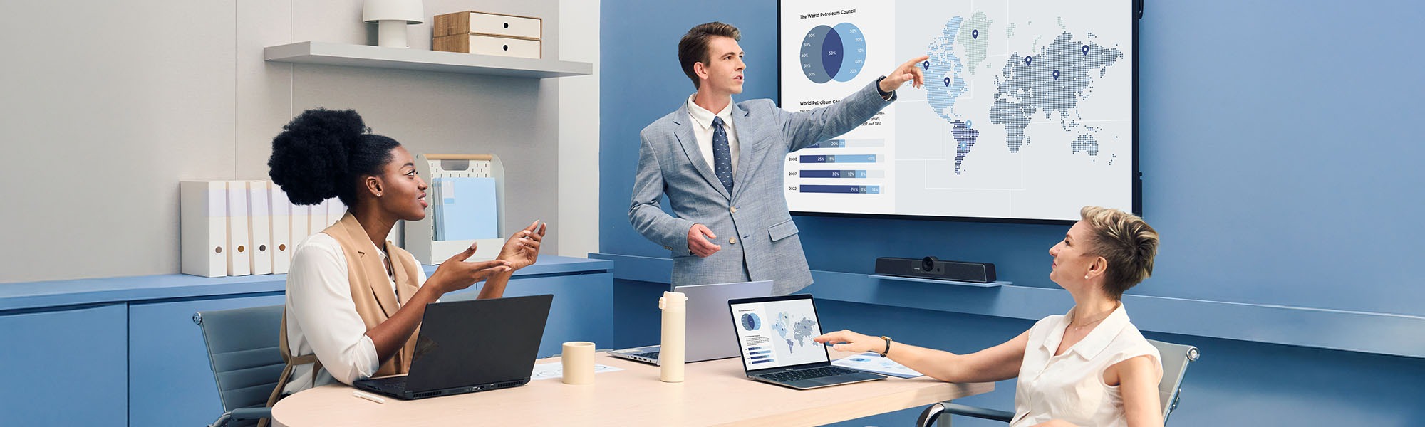 man standing and pointing to a CDE31 display with 2 women coworkers sitting at a table with laptops also looking at the CDE31
