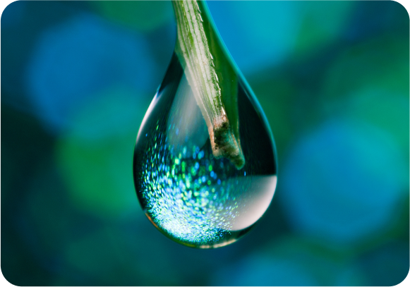 A close-up of a water droplet hanging from a green stem, with a soft blue-green bokeh background. The droplet reflects light, creating a sparkling effect.